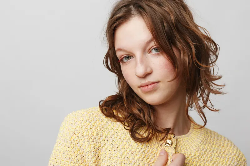 Young woman with curly brown hair wearing yellow sweater looks thoughtfully at camera on white background. Concept of natural beauty, casual style, and soft lighting portrait.