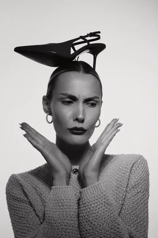 Slim white woman with pale skin and slicked hair poses with black high heel shoe on her head, wearing knitted sweater and hoop earrings in black and white studio portrait.