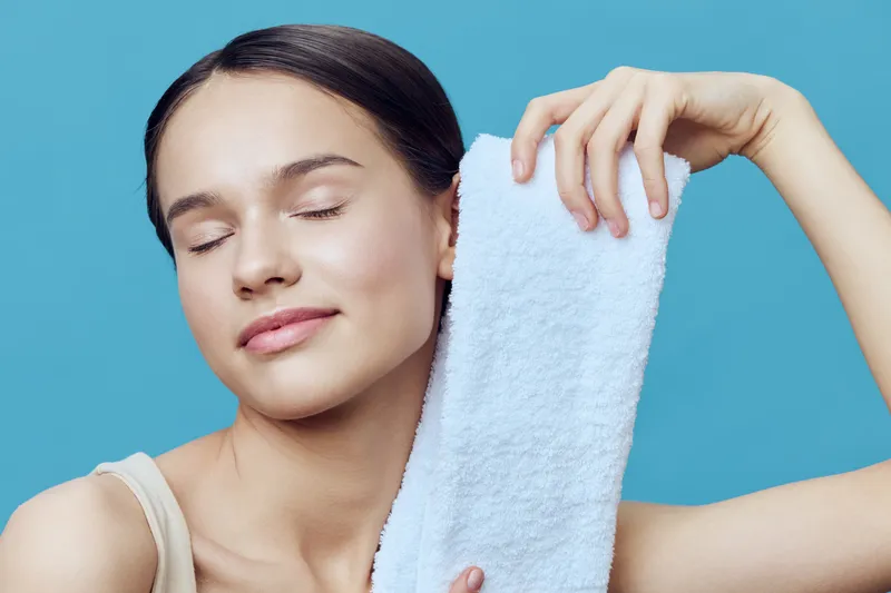 Young woman with closed eyes enjoying a towel on her shoulder, representing relaxation and self care in a serene turquoise background