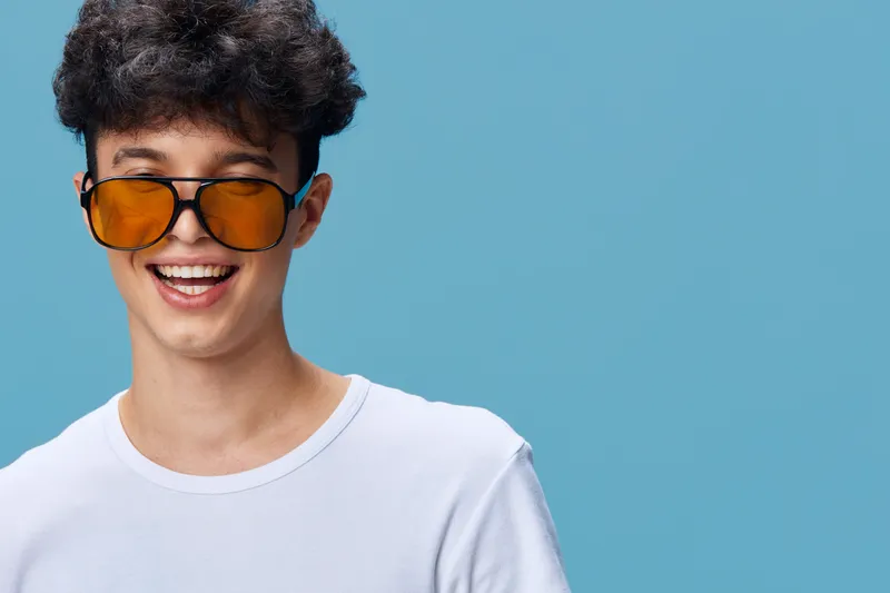 Young man smiling wearing oversized orange sunglasses and white t-shirt, curly hair, bright blue background, cheerful urban casual style with vibrant colors.