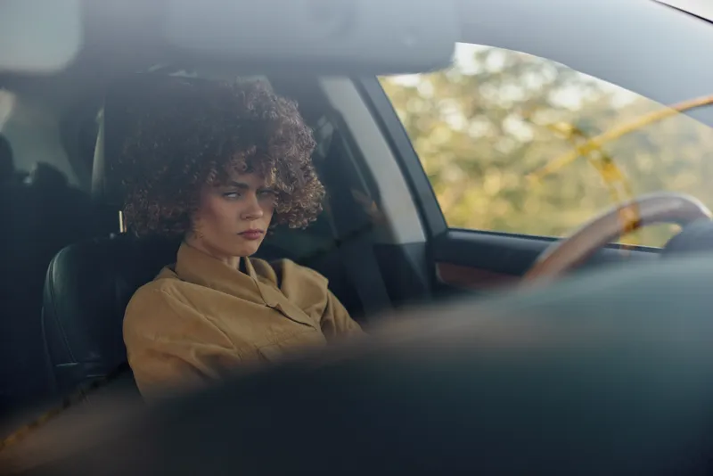 Woman sits in car interior with contemplative expression, vintage film color palette, lifestyle moment captured behind the wheel, calm mood, everyday commute scene, warm cinematic atmosphere