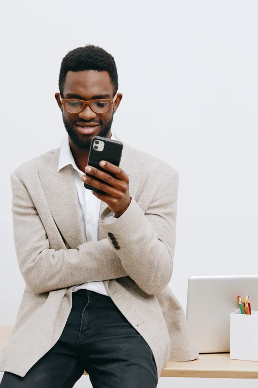 Professional man using smartphone in office setting, wearing glasses and casual beige sweater, sitting near a desk with a white background, focused on device. People lifestyle concept, modern technolo