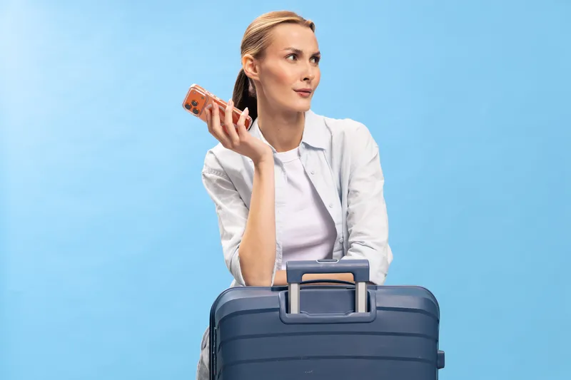 Young woman in white shirt holds smartphone while leaning on blue suitcase against light blue background. Concept of travel, planning, and vacation preparation with modern technology.