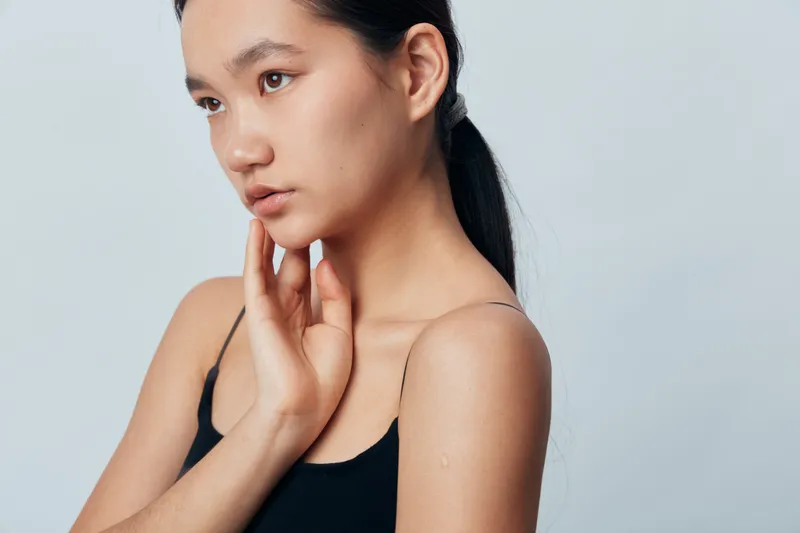 Young Asian woman in black sleeveless top with natural makeup and ponytail hairstyle looking sideways with hand on neck on plain light background. Portrait, beauty and calm expression.