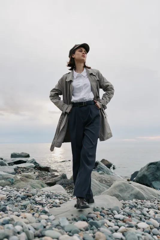 Young woman stands confidently on rocky beach wearing casual jacket and cap. Overcast sky and calm sea create moody atmosphere, concept for fashion, confidence, and outdoor lifestyle.