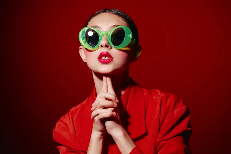 Young fashionable woman with bright red lipstick wearing oversized green sunglasses and a red jacket. Bold creative look with dramatic lighting on a red background.