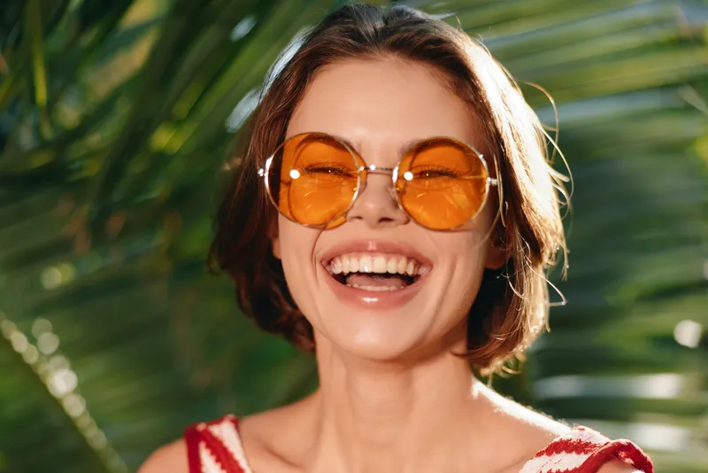 A cheerful woman wearing orange sunglasses smiles broadly in a sunny outdoor setting, capturing a carefree, vibrant mood and a sense of sunshine, warmth, and positivity.