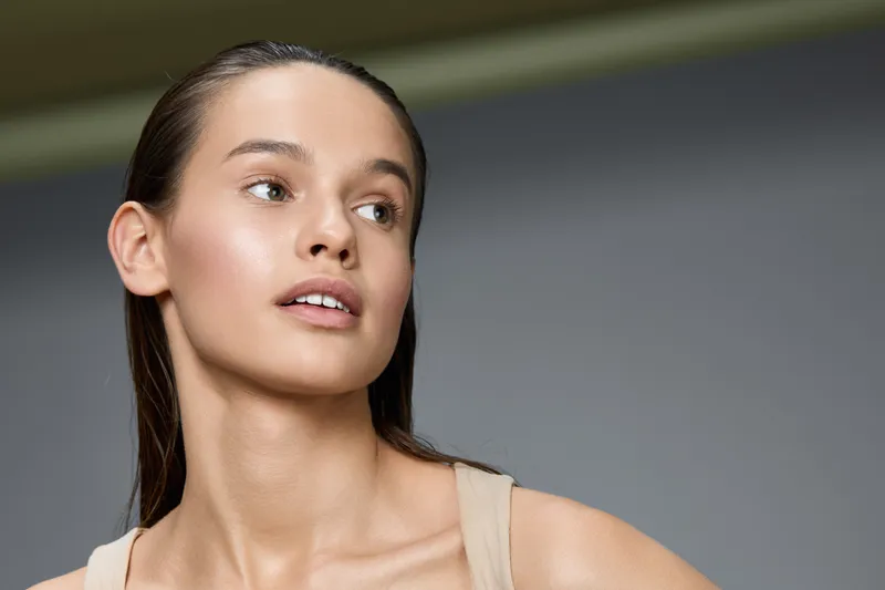 Young woman with glowing skin in a neutral tank top, showcasing a natural beauty look against a soft gray background, embodying wellness and self care