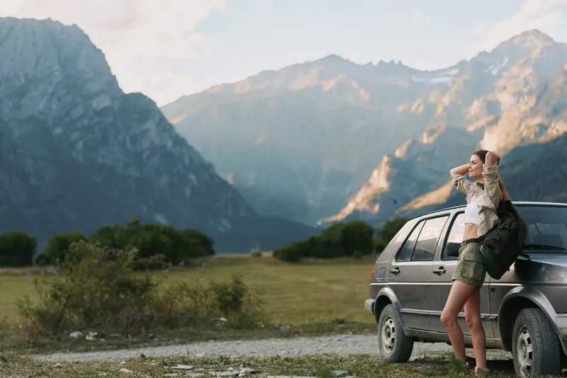 A woman by a car gazes toward distant mountains during golden hour, capturing a travel lifestyle moment of outdoor exploration, freedom, and calm.