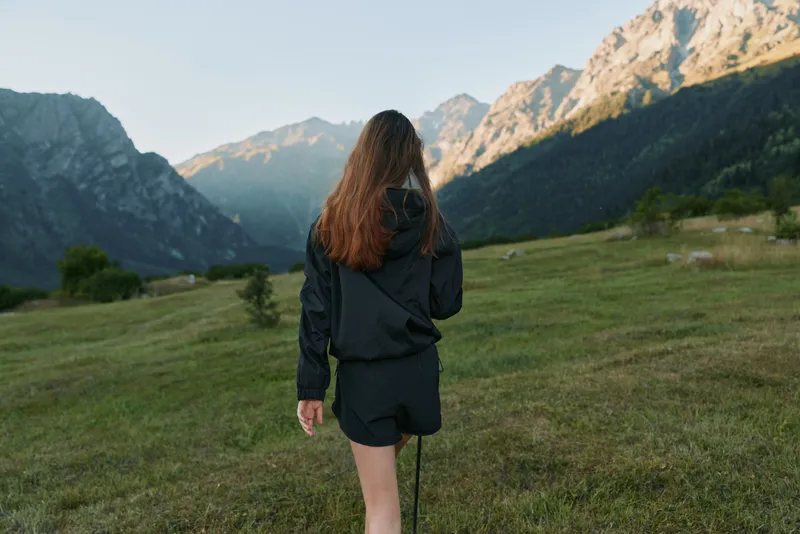 A lone hiker walks through a lush alpine meadow, seen from behind. Dressed in a dark jacket and shorts, she explores vast mountains, exuding serenity and curiosity in a spacious outdoor setting.