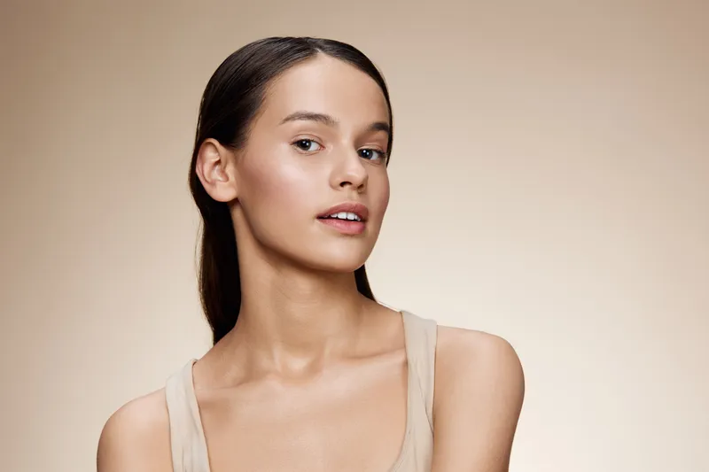 Natural beauty portrait of a young woman with smooth skin and a soft expression against a warm beige background, embodying confidence and simplicity in beauty