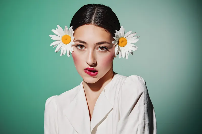 Young woman with bright makeup wearing white clothes and two large flowers in her hair poses against a soft green background. Creative beauty and fashion concept with fresh style.