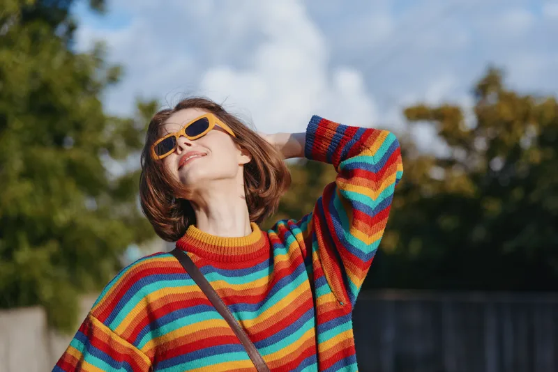 Woman in a rainbow sweater wearing yellow sunglasses, smiling with short hair outdoors in a park, casual portrait showing relaxed pose and hand in hair, lifestyle fashion travel vibe