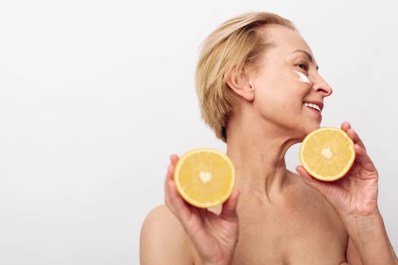 Mature woman with short blonde hair holds two halves of orange near face, applying white cream under eyes. Concept of natural skincare, vitamin c, healthy lifestyle, and facial beauty care.
