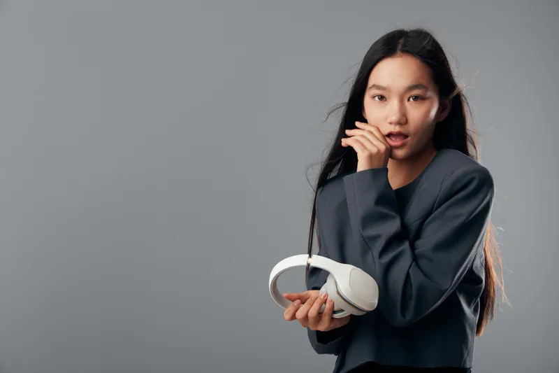 Young woman with long hair holding white headphones with thoughtful expression against gray background. Female portrait, casual wear and technology accessory for music listening or podcast.