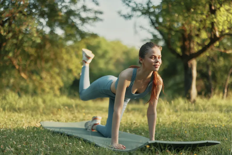 Young woman practicing yoga outdoors on a green mat in a park, promoting health and wellness, wearing activewear with serene expression and vibrant nature background