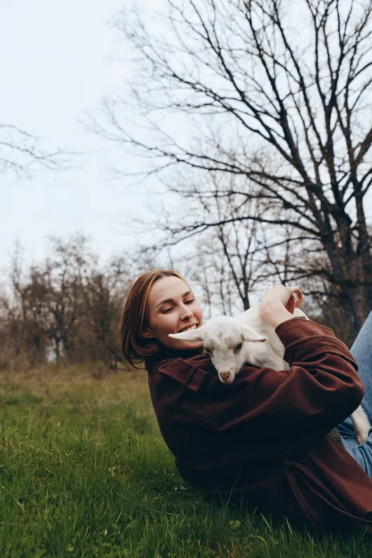 Smiling woman relaxing on grass, holding a playful goat kid in a cozy brown jacket. Outdoor lifestyle scene showing companionship, nature, and gentle care. Soft sunlight creates a warm, happy mood.
