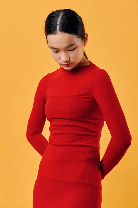 Young woman wearing red dress standing with hands behind back and looking down on yellow background. Female model posing in studio with serious expression and long black hair.