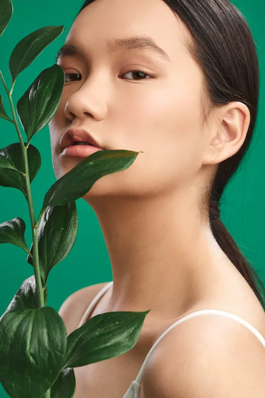 Asian woman with delicate features poses beside green leaves, creating a serene portrait that emphasizes natural beauty and calm studio aesthetics.
