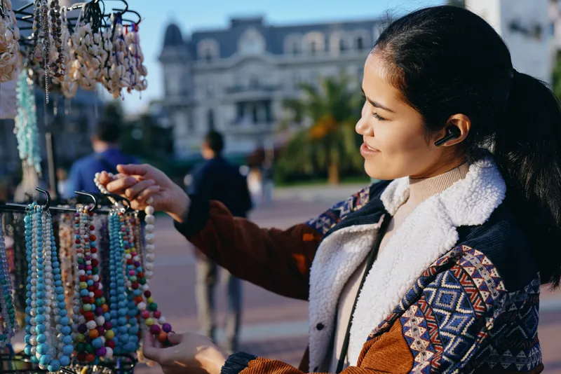 Colorful beads shimmer as a market shopper browses a wooden stall, selecting a handmade necklace and accessories under bright daytime sunlight, capturing artisan craft and urban style