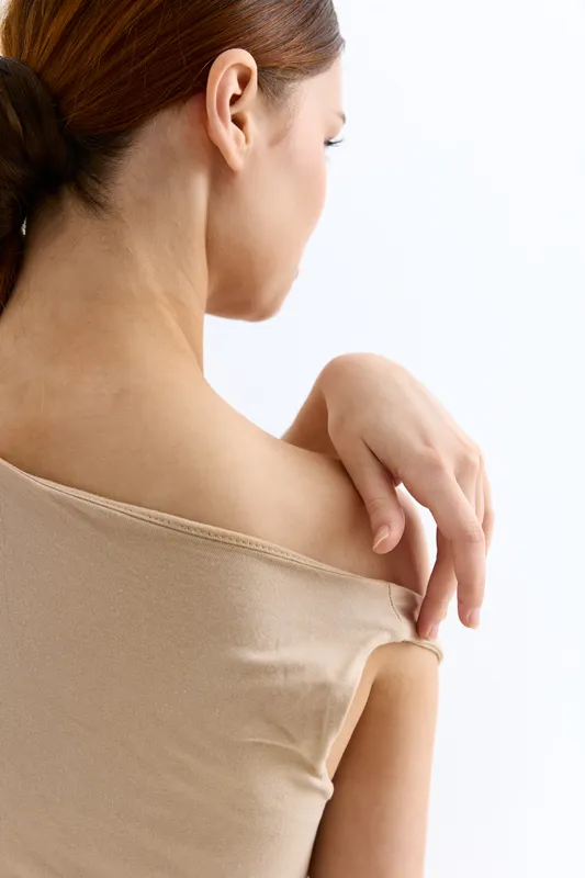 Young woman with bare shoulder gently touching skin, wearing beige off-shoulder top against plain white background, soft natural light, delicate feminine pose.