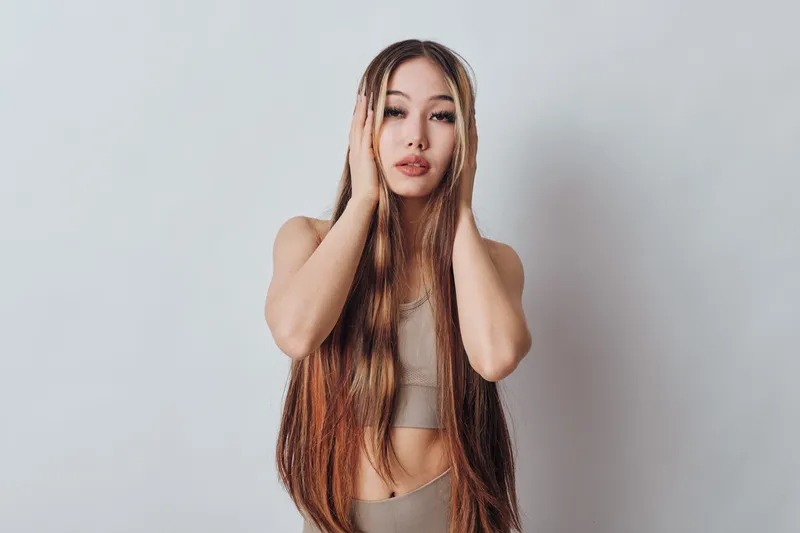 Young woman with long braided hair posing with hands on head against plain background. Female model with natural makeup in casual outfit showing thoughtful expression.