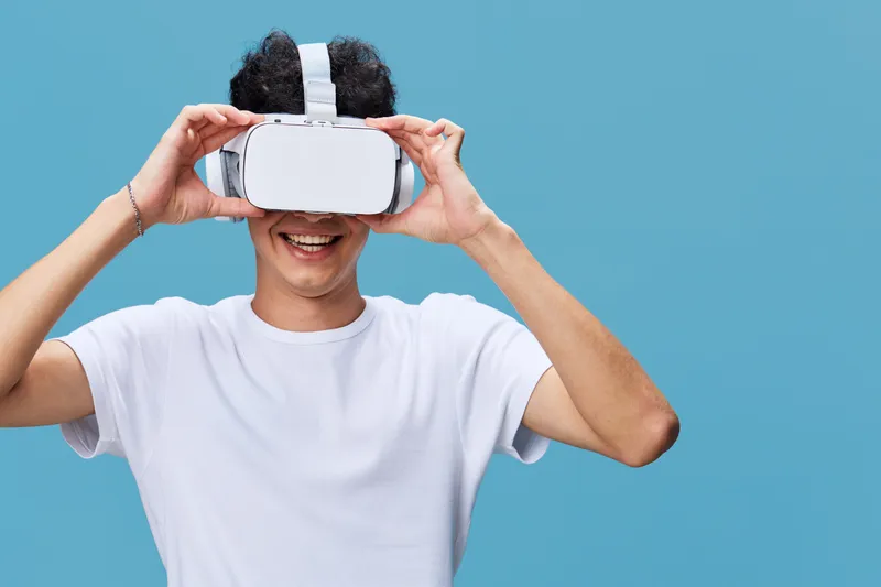 Young man wearing virtual reality headset enjoying immersive digital experience, smiling and relaxed against blue background in casual white t-shirt.