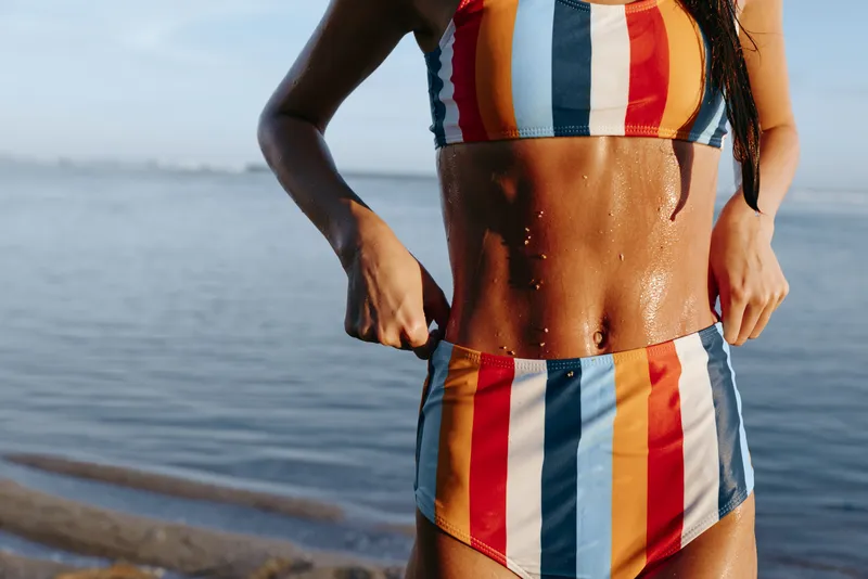 Woman in striped bikini on seaside ocean in summer, close up toned torso with confident posture, outdoor fitness lifestyle concept, sunlit warm pier background, gentle waves