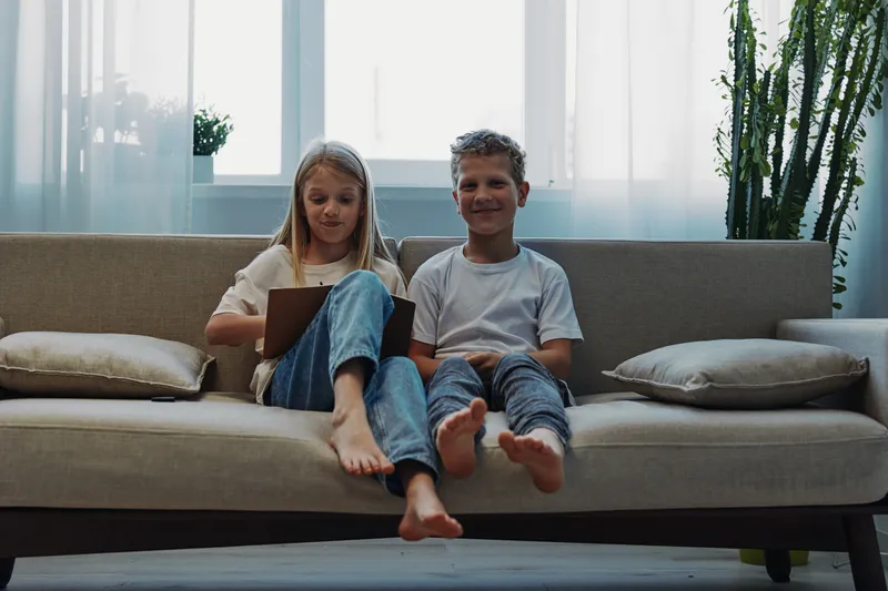 Two children sit barefoot on a beige sofa in a bright living room, a girl focused on a tablet and a boy smiling. Concept of childhood, technology, home comfort, and sibling bond.