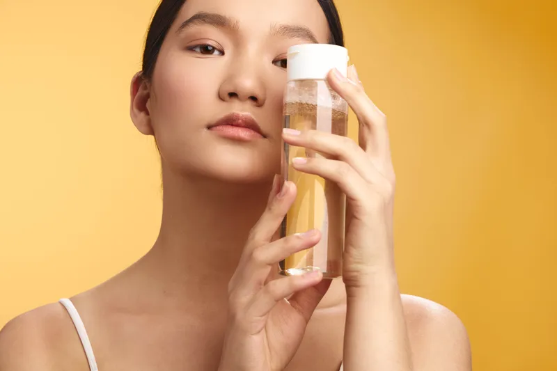 Asian woman stands with a clear bottle in a soft studio setting, posed against a warm yellow background. She gazes calmly, conveying purity, freshness, and a subtle lifestyle moment.