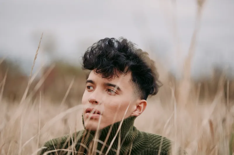 A young man with curly hair sits among tall grasses in a muted field, wearing a green knit sweater, conveying a quiet, authentic presence and focus.