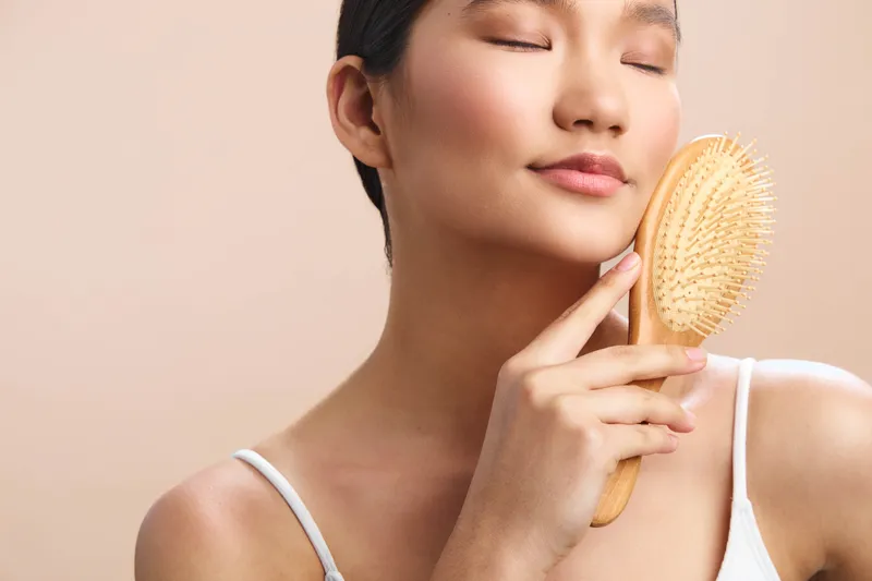Asian woman with closed eyes smiles softly as she uses a wooden body brush against her shoulder, creating a calm, intimate skincare moment in a neutral studio.