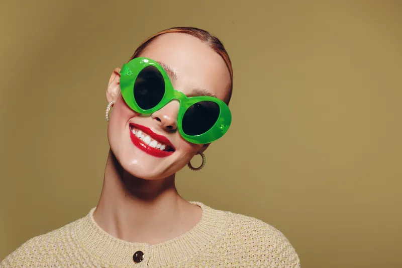 Smiling woman with slicked hair wears large green sunglasses and red lipstick. She poses against a plain beige background wearing a beige knitted sweater. Concept of fashion and style.
