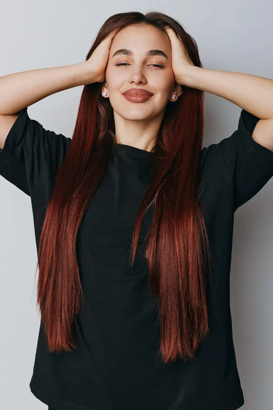 Woman with long red hair and closed eyes smiling with hands on head wearing black shirt standing against white background. Relaxation, happiness and calm mood in casual style portrait.