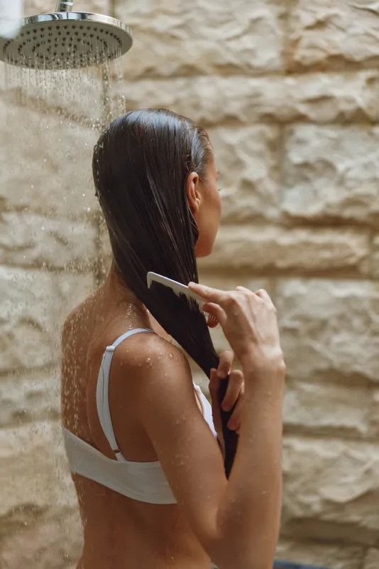 woman combing wet hair under outdoor shower with stone wall background, natural light, summer wellness moment, healthy lifestyle concept