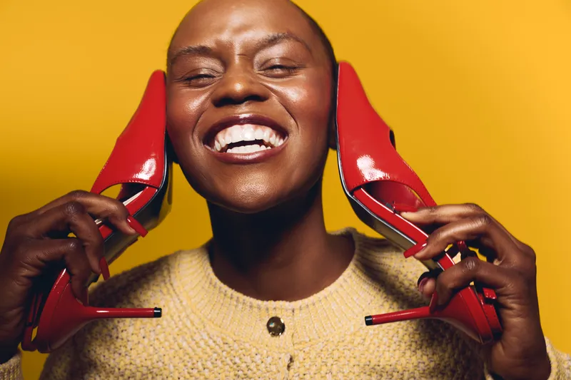 Happy African woman with dark skin holds red high heels near her face smiling broadly against yellow background. Concept of joy, fashion, beauty, and confident black female model.