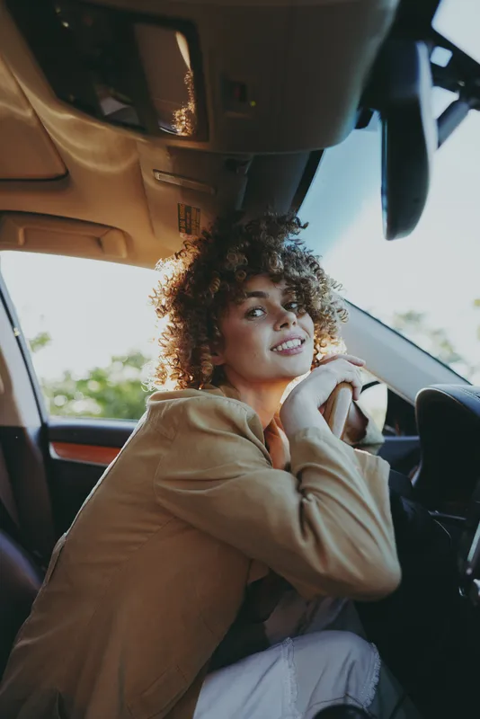 Young woman sits in a car interior wearing a casual hoodie, warm daylight filters through the window, a relaxed lifestyle portrait with a friendly candid mood