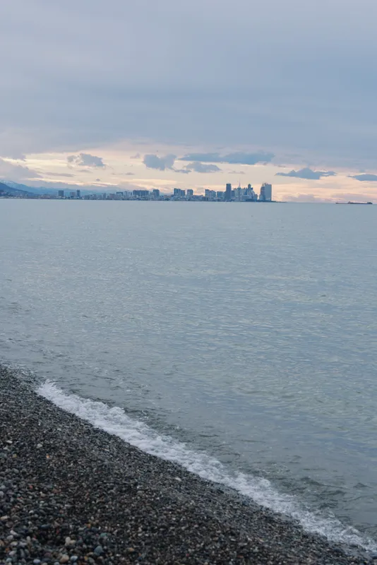 Coastal scene with calm water, pebbled beach in the foreground and distant city skyline under soft clouds, tranquil blue tones suited for travel, nature and urban waterfront themes