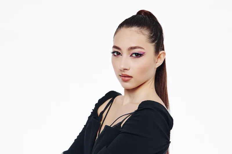 Young woman with dramatic pink winged eyeliner and glossy lips wears a trendy off-shoulder black top on white background. Concept of beauty creative and fashion styling.