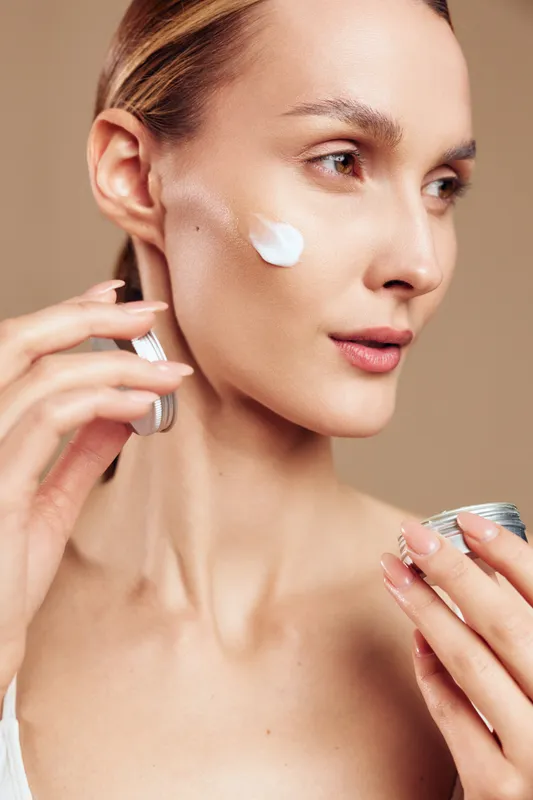 Closeup of young woman applying moisturizing cream on face with natural skin tone holding cosmetic jar, skincare routine, beauty concept on beige background with gentle light and calm expression