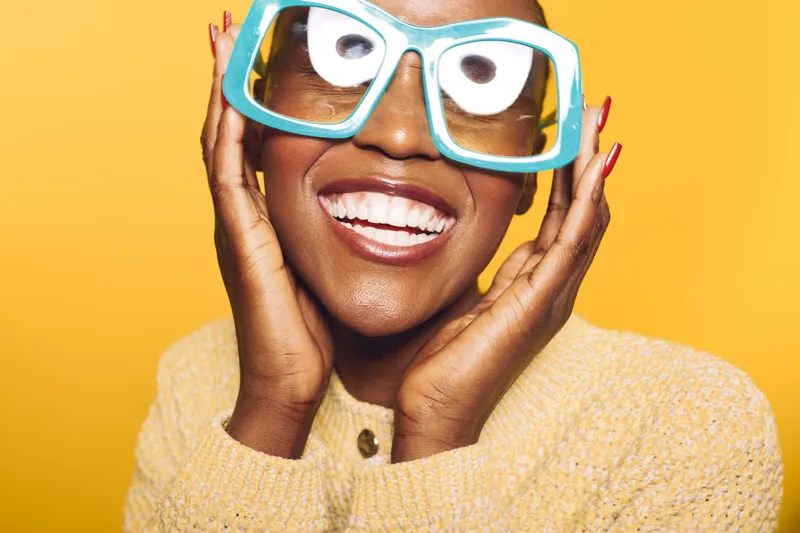 Happy African woman with dark skin wearing large blue glasses and a yellow sweater smiles joyfully on a bright yellow background. Concept for happiness, fashion, and beauty.