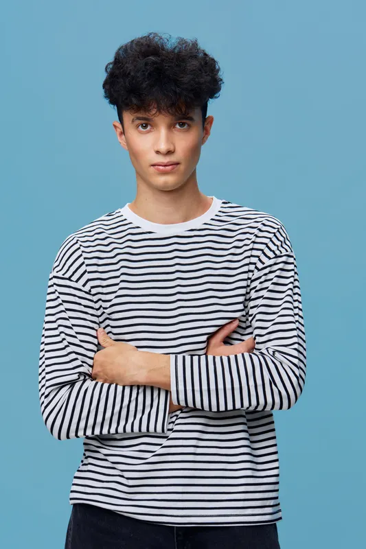 young man with curly hair wearing striped shirt standing with crossed arms against solid blue background, casual style, confident expression, studio portrait