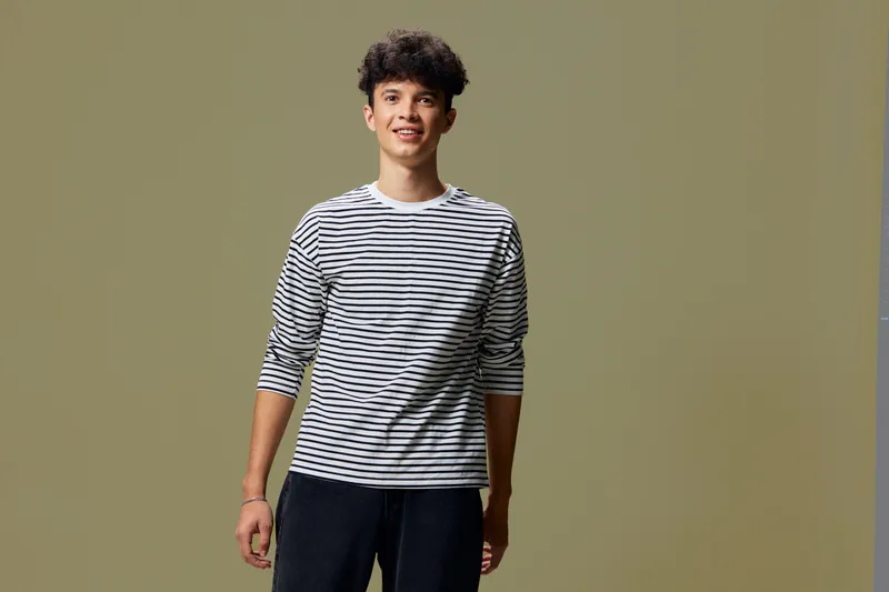 Young man wearing casual striped shirt stands smiling against olive green background, displaying relaxed and friendly expression in studio lighting.