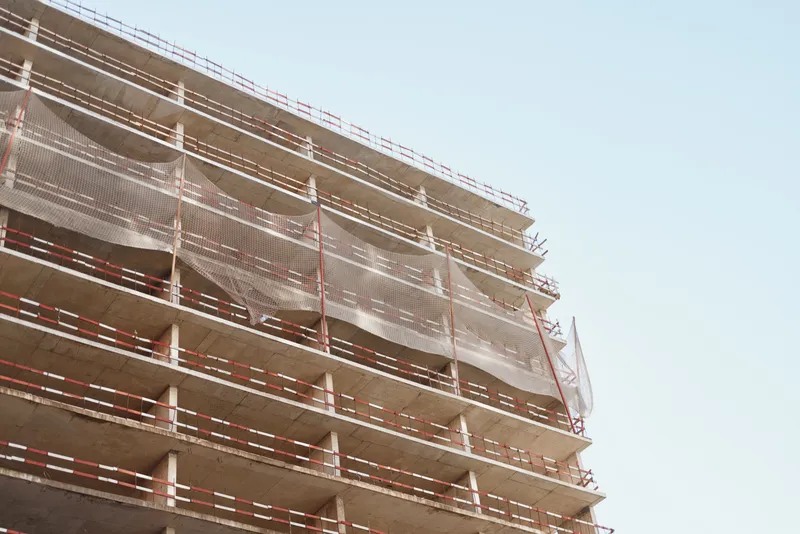 Unfinished multi-story concrete building stands under clear blue sky with protective netting and scaffolding visible along its edges during daytime construction.