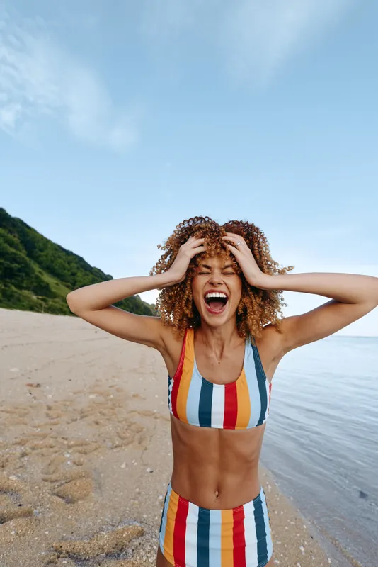 Woman, beach, laughing, striped swimsuit, outdoors, summer sunlight, hands on head, ocean view, green hills, warm colors