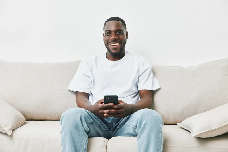 Smiling African American man sitting on a beige couch, holding a smartphone, dressed casually in a white t shirt and jeans, exuding happiness and relaxation