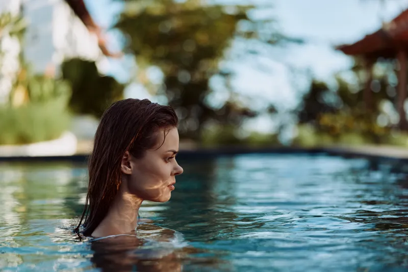 Serene woman with wet hair in a pool, side profile, enjoying a sunny day. Water reflections glow on blue surface, outdoor setting, casual wellness and leisure. Calm mood, fresh air, self care through