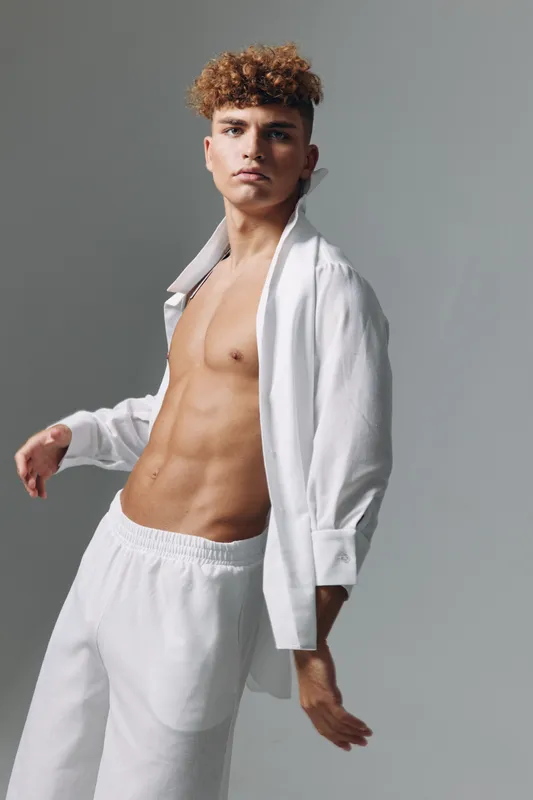 Young man with curly hair posing in white open shirt and pants against gray background. Fashion model with toned body and confident expression in studio lighting.