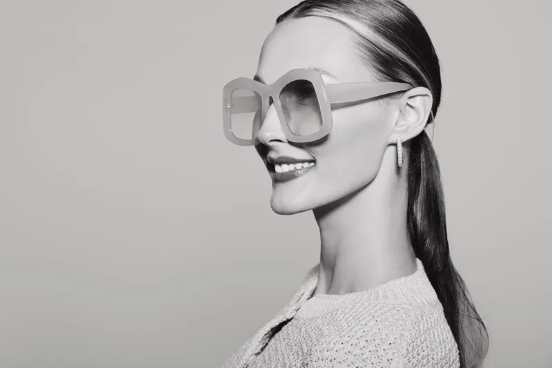 Side portrait of a smiling Caucasian woman with pale skin wearing large sunglasses and a textured sweater in a studio. Fashion and style concept.