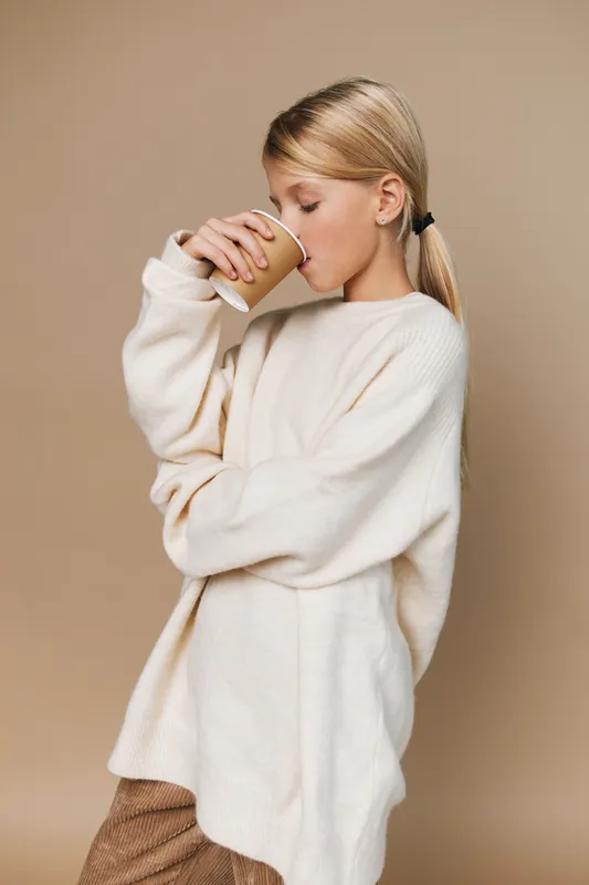 Young woman sips from a paper cup, wearing an oversized cream sweater against a warm beige backdrop. Cozy, relaxed moment that conveys comfort, ease, and quiet everyday routine.